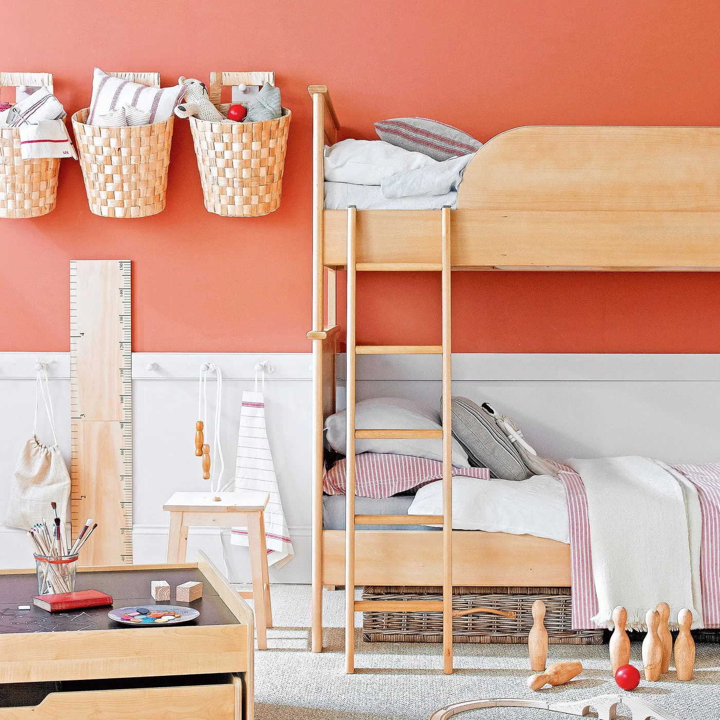 A plain wood bunk bed stands against a coral wall with baskets above, white storage below and a small wood desk in front