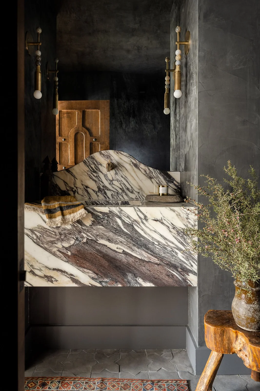 A broad pale dark-veined basin counter and shaped backsplash sit above plain dark drawers against charcoal walls beside slim hanging lights and a small wood stool
