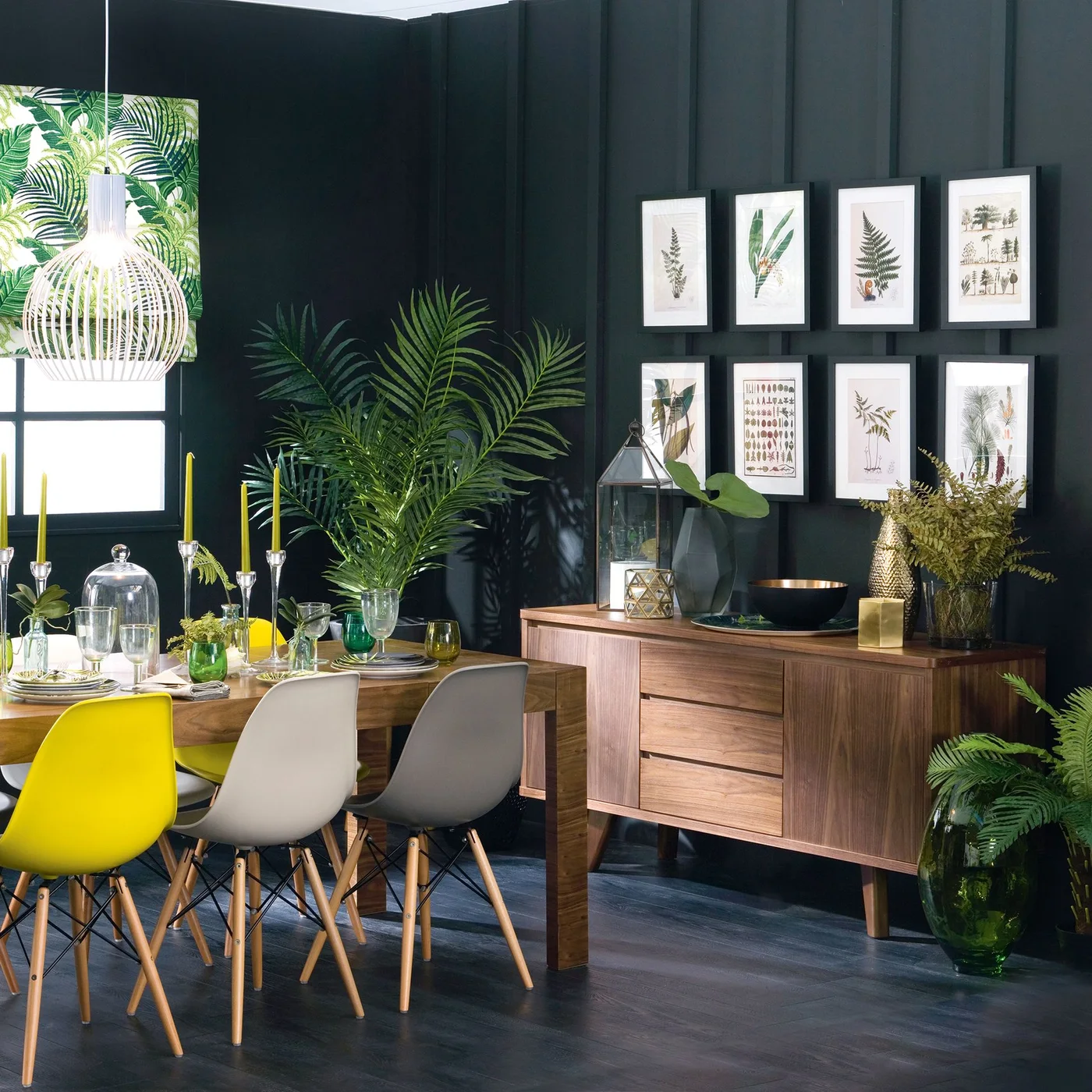 A wood sideboard with drawers and a cupboard stands beneath botanical pictures beside a dark dining-room wall, plants and mixed-color chairs