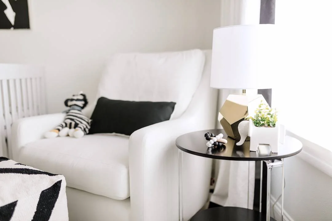 A white nursery chair with a dark cushion sits beside a round dark table, white-shaded lamp and small decorative objects