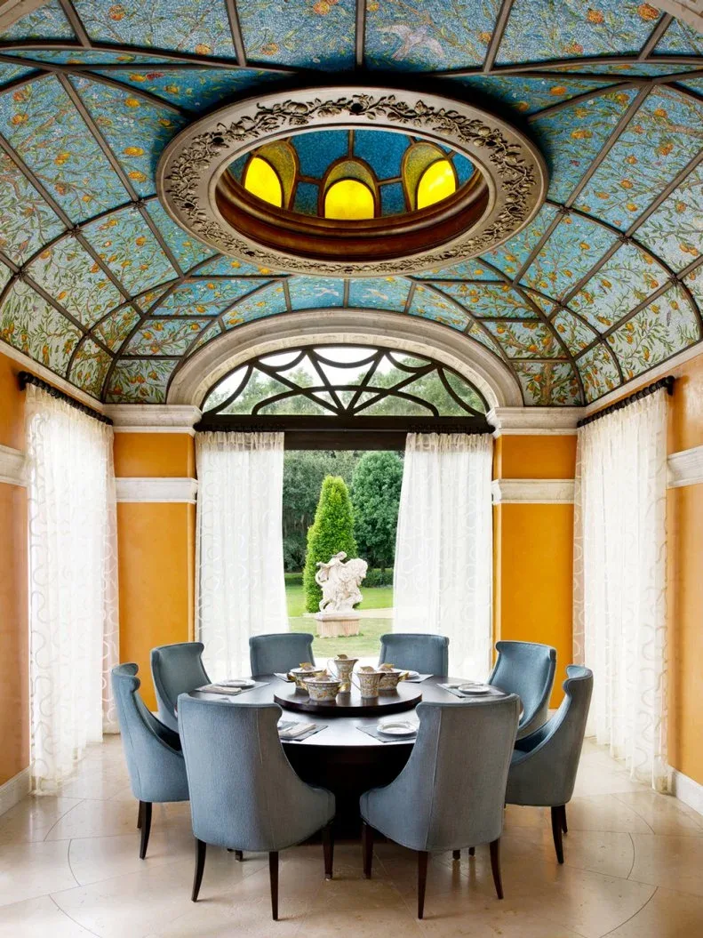 Pale blue upholstered dining chairs with tall curved backs around a dark round table, beneath a patterned blue ceiling and beside orange walls.