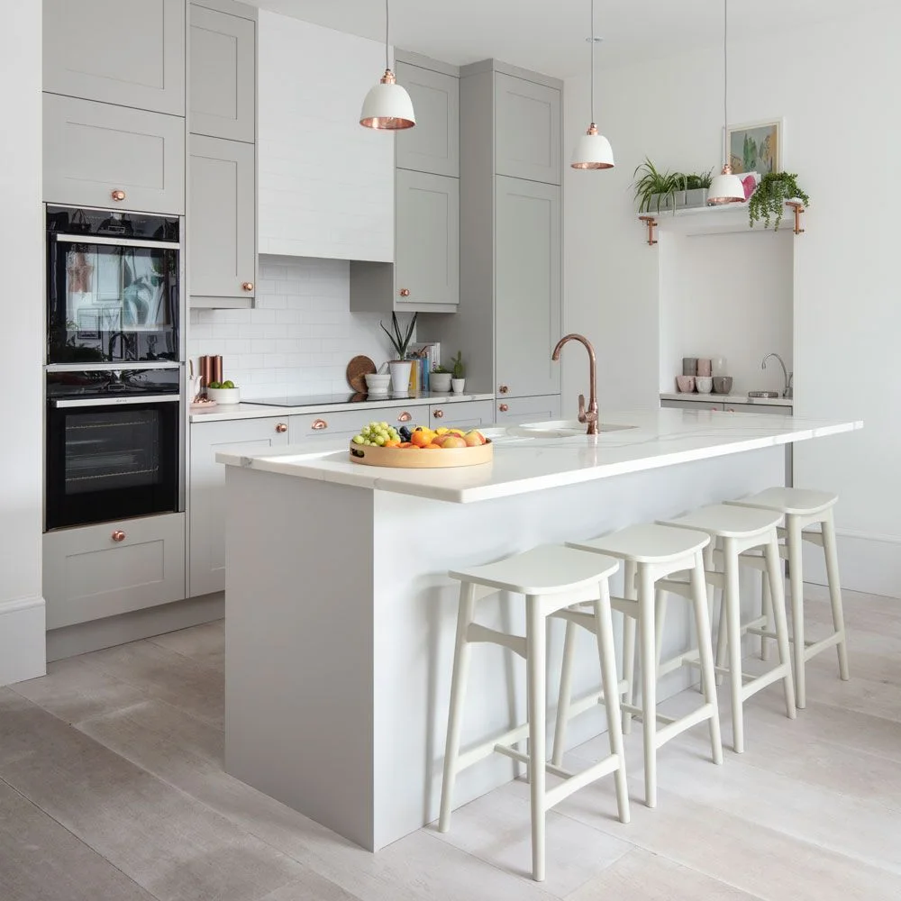 Tall pale gray cabinets and an oven housing stand behind a white island with stools, copper-toned faucet and small pendant lights