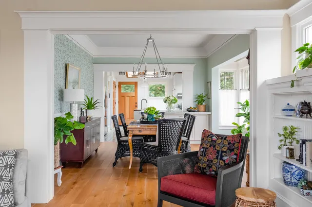 Timber dining table with dark patterned chairs beyond a gray armchair with a red seat and floral cushion