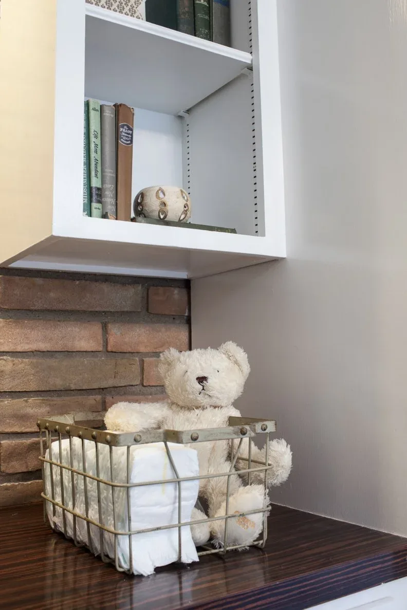 Wire basket with folded supplies and soft toy beneath pale shelves beside brick-textured wall.