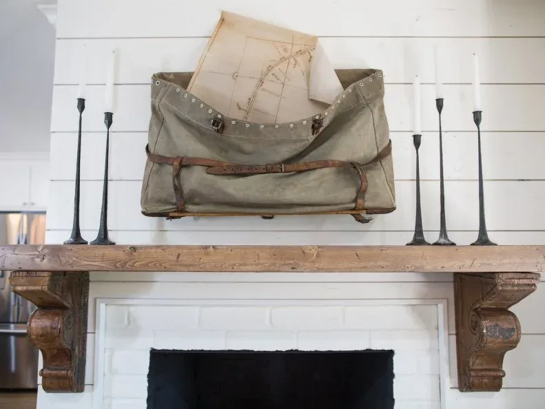 Wood mantel on decorative corbels above pale brick opening, with tall dark candlesticks and a hanging fabric bag containing papers