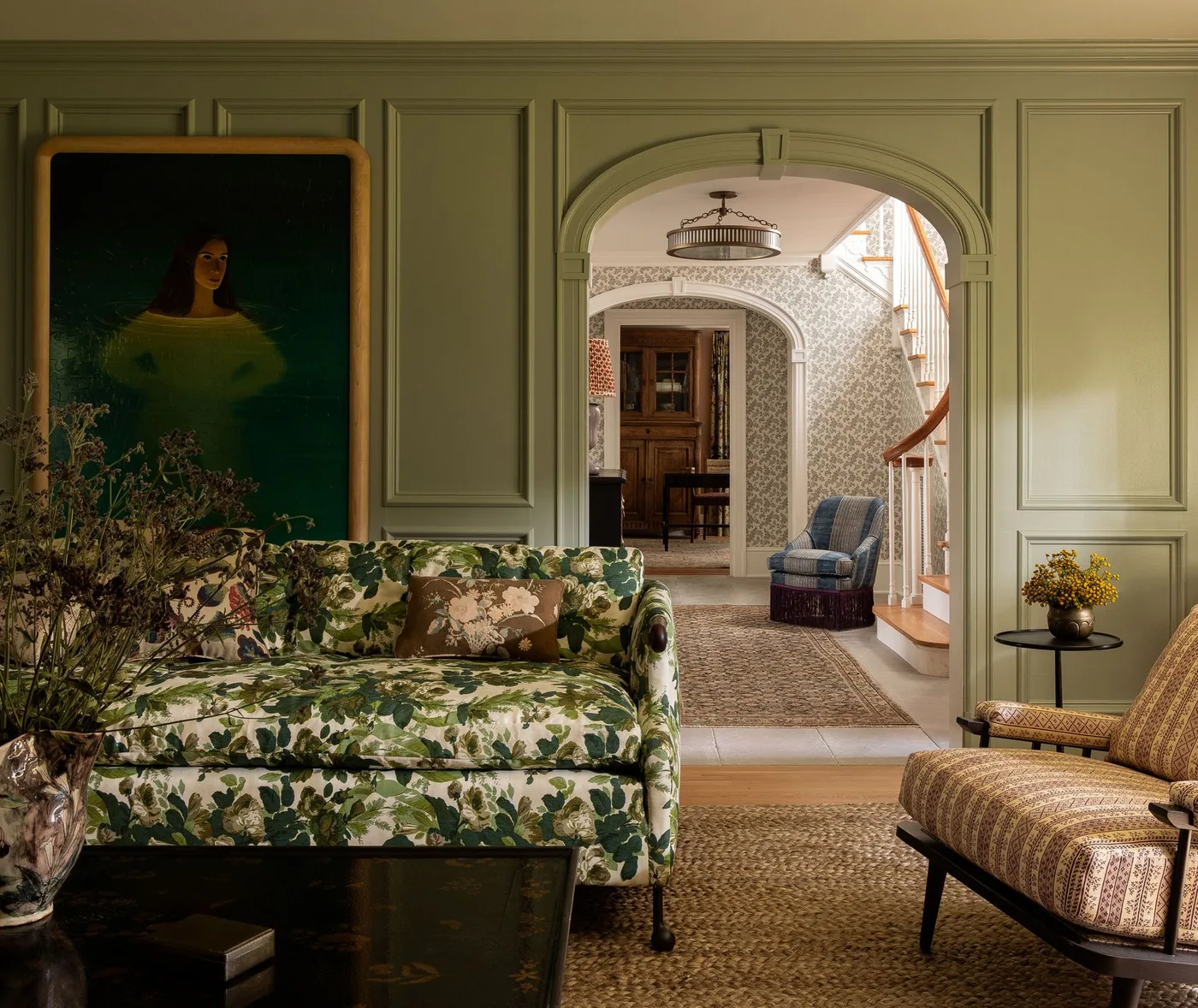 A green-and-pale botanical sofa sits beside patterned seating against muted green paneling near a dark round table