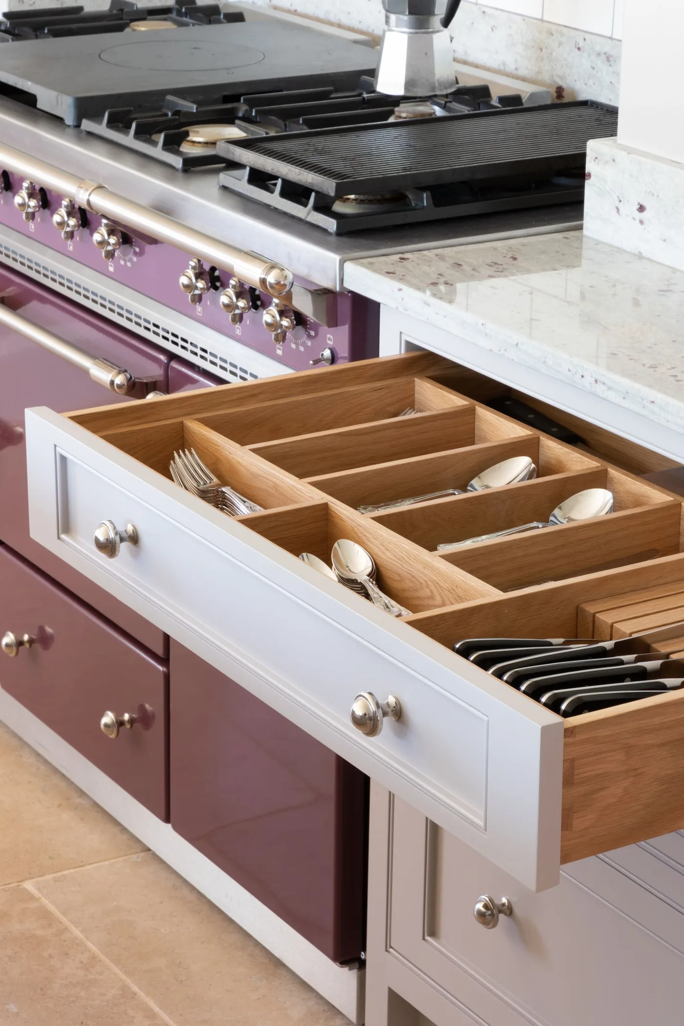 Country kitchen with pale open drawer and wooden cutlery dividers beside a purple cooker and pale counter.