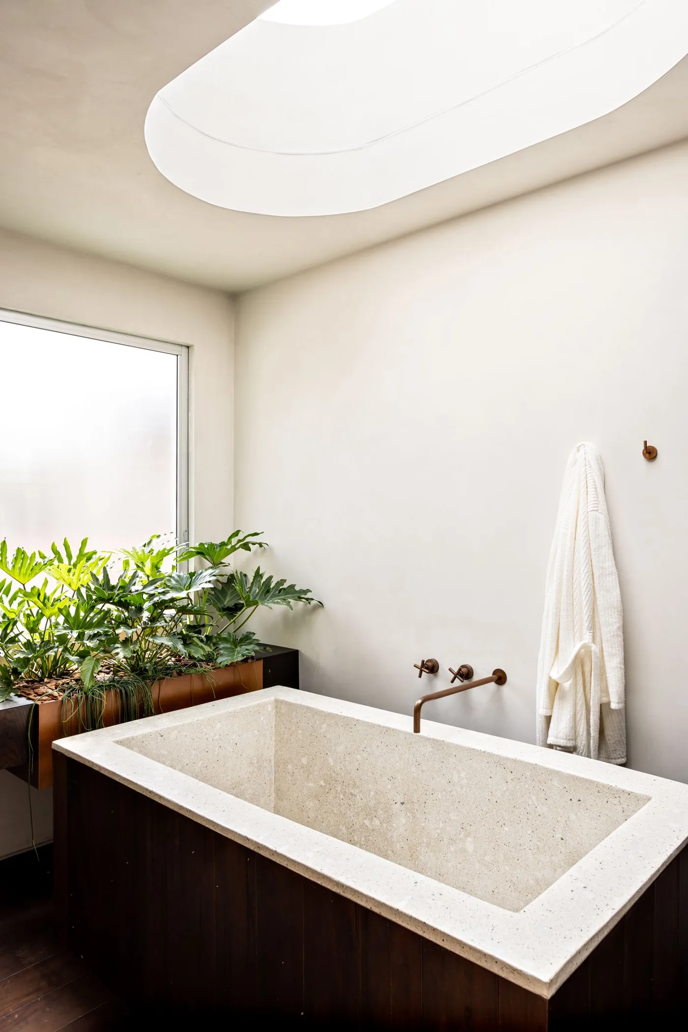 Pale speckled rectangular bath beneath an oval skylight with greenery by a frosted window