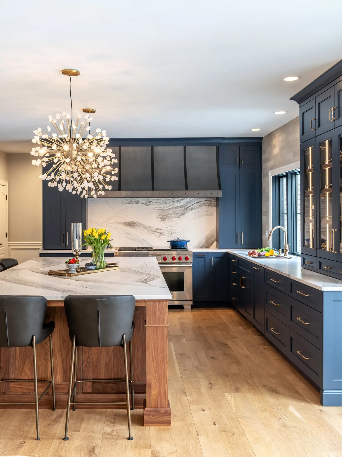 Deep blue kitchen cabinets surround a timber-front island with pale veined counter beneath a decorative pendant beside glazed storage and windows