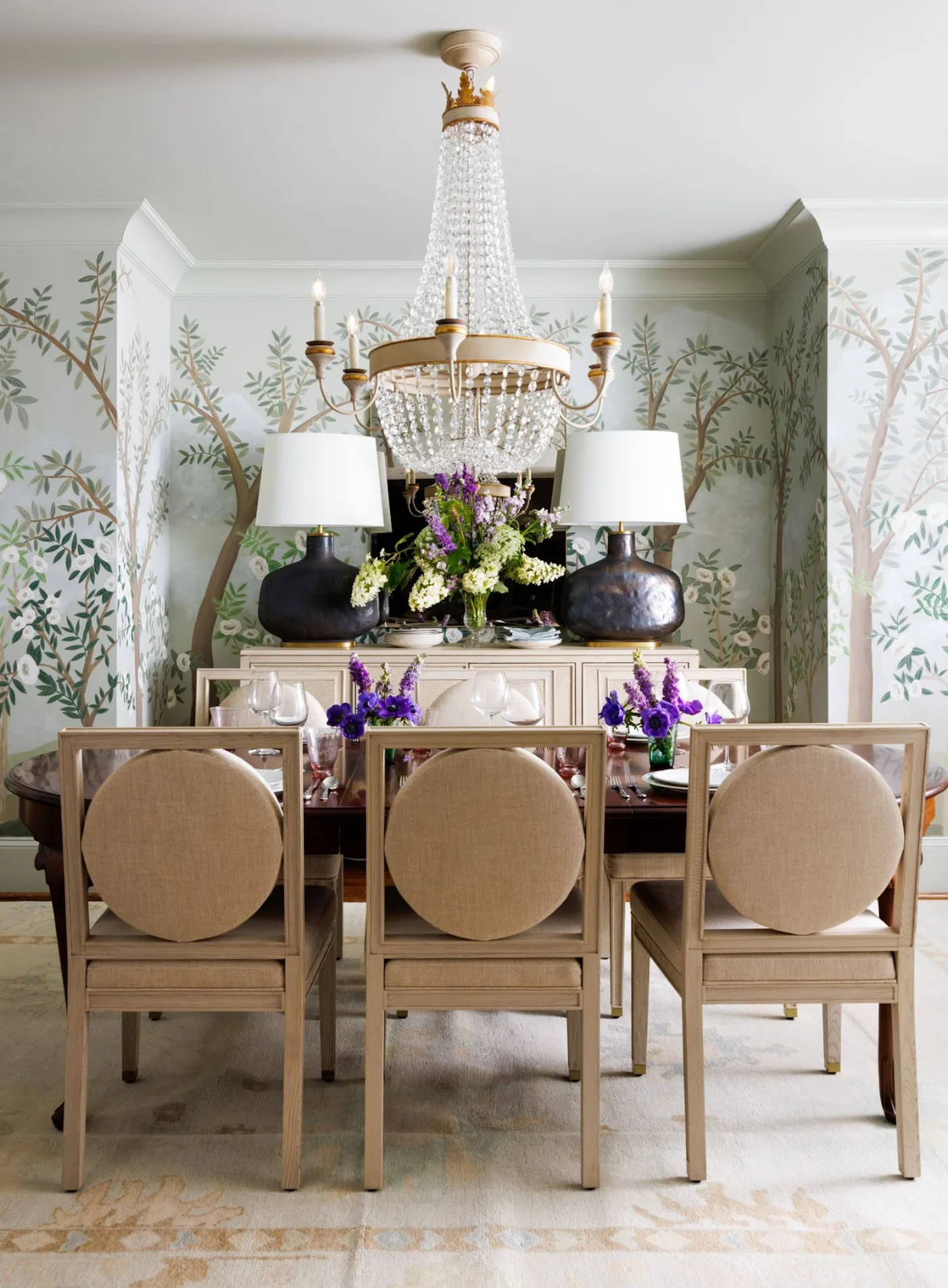 Round-backed dining chairs beneath a clear chandelier with tree-patterned walls and a pale sideboard.