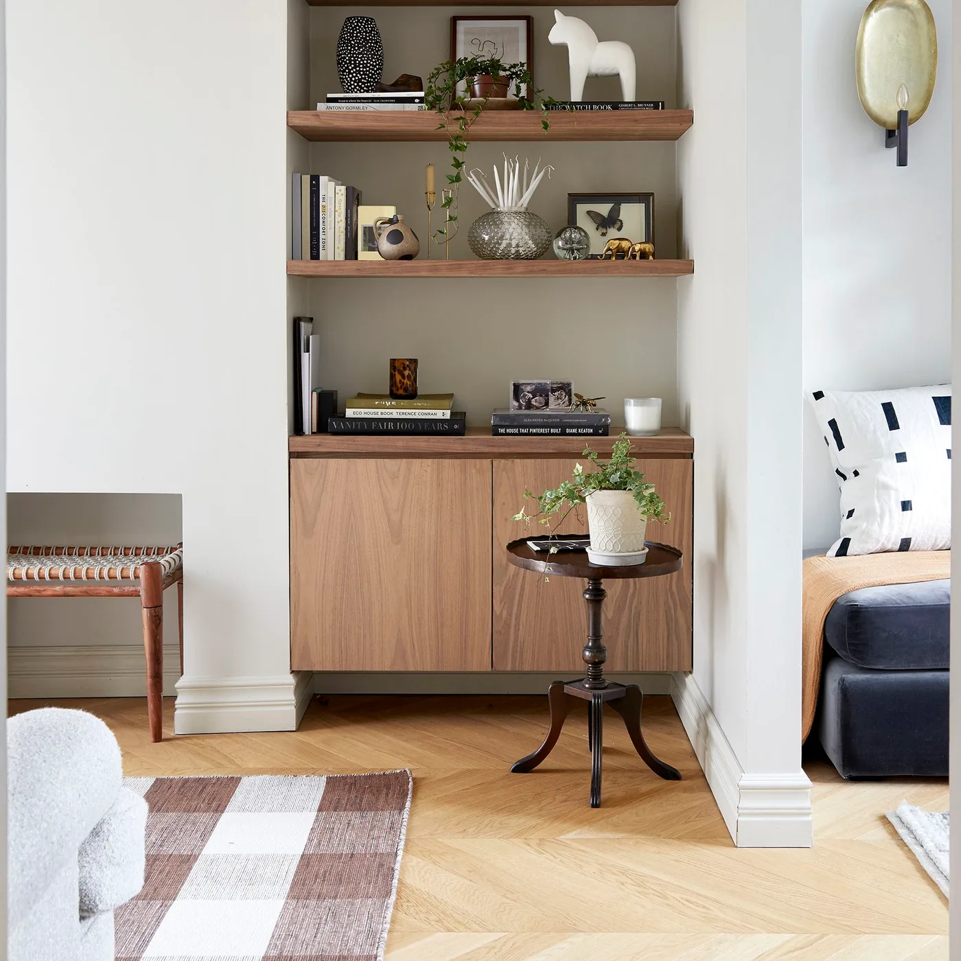 Wood shelves and a flat-front lower cabinet fit into a pale wall recess beside a small dark round table, light flooring and a checked rug