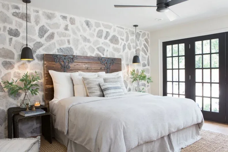 White bedroom with irregular gray stone-looking bed wall, wood headboard and black glazed doors