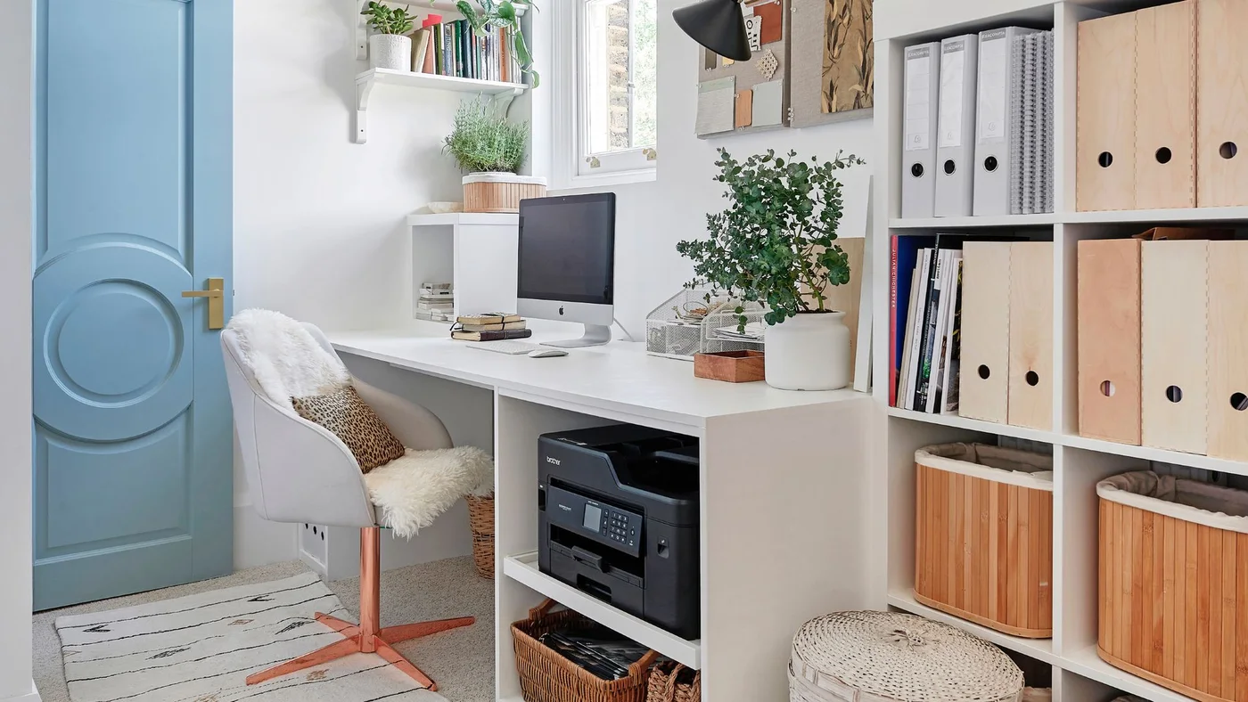 A long white desk and cubby shelves surround a gray upholstered chair near a blue door, with files, baskets and a printer stored below