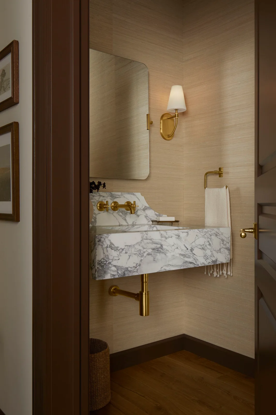 A pale dark-veined wall basin sits beneath a rounded rectangular mirror against warm pale walls beside a small shaded wall light and a white towel