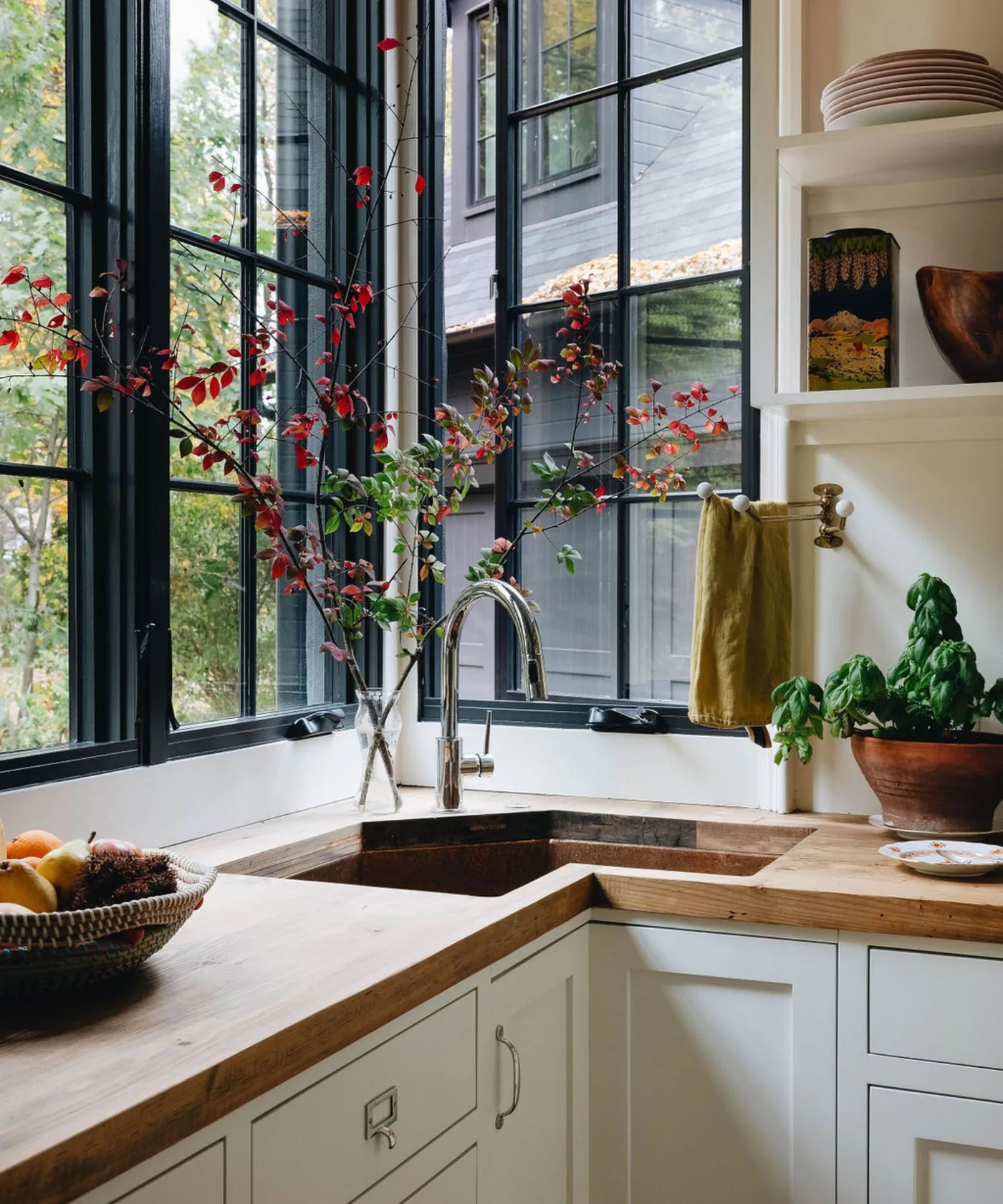 Cottage sink corner with pale framed cabinets, wooden countertop, dark divided windows, tall faucet, open shelves and a small potted plant.
