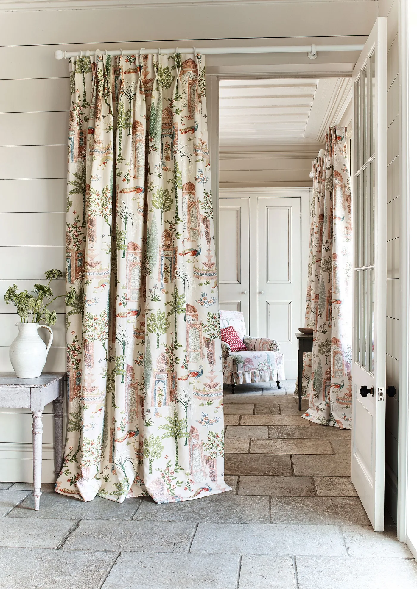 Country hallway with pale horizontal paneling, botanical curtains across an opening, a narrow painted side table and large rectangular floor tiles.