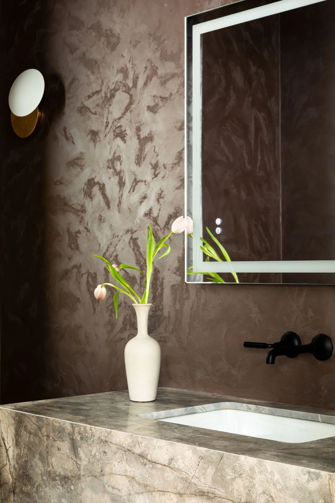 A pale veined basin counter sits against a warm brown textured wall beneath a rectangular mirror beside a small curved light and black wall-mounted taps