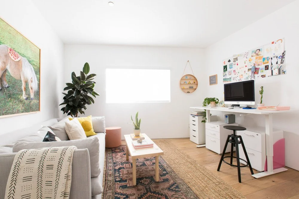A pale gray sofa sits opposite a white desk and drawer unit in a bright living space with plants, a patterned rug and a black stool
