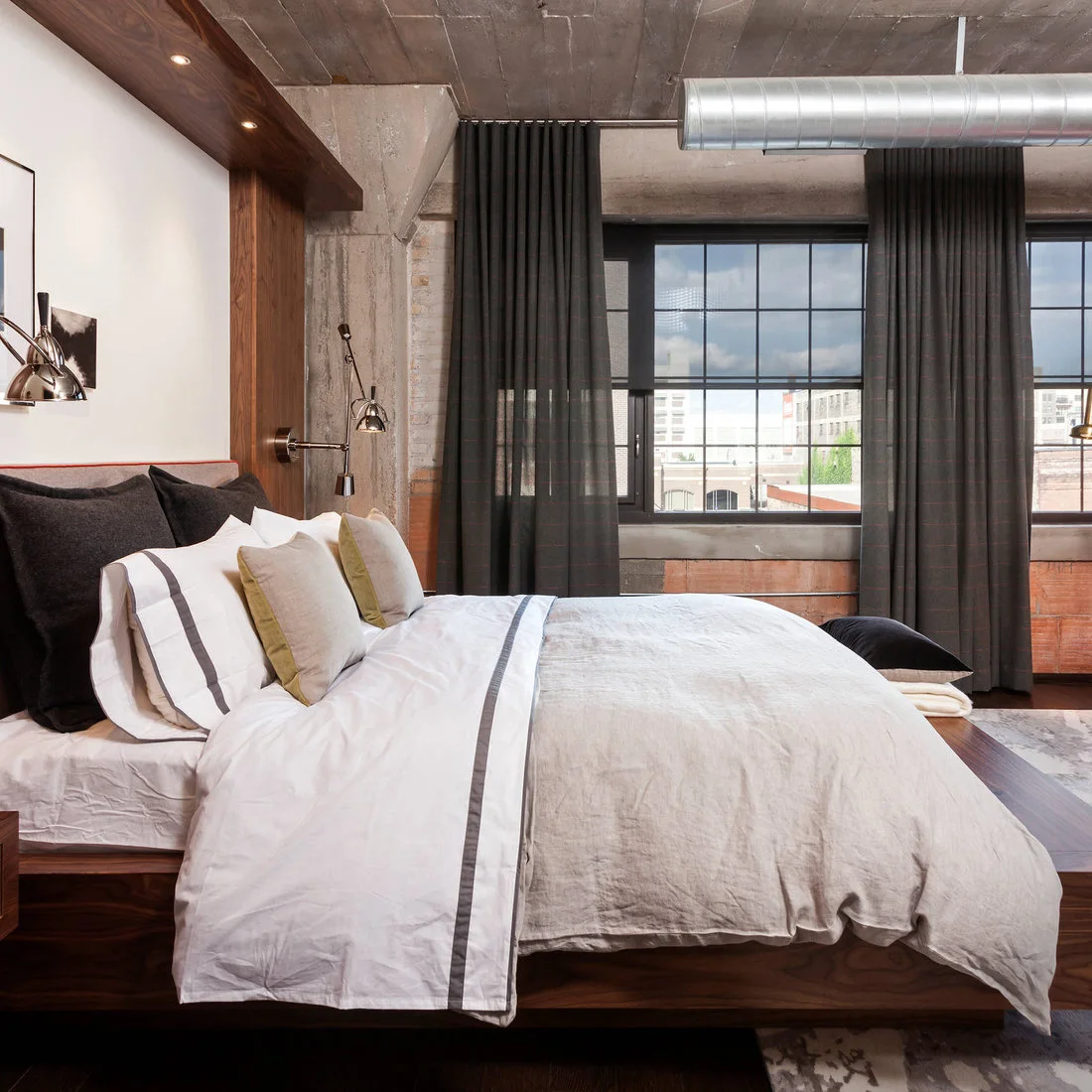A wooden bed with pale striped bedding sits beside dark curtains and a grid window beneath exposed silver ductwork and a rough ceiling