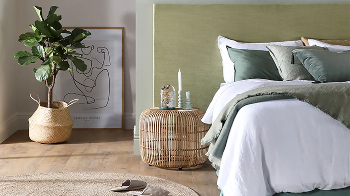 A pale bed with green cushions and a throw sits against a muted green wall beside a rounded woven table, leafy plant and framed line drawing
