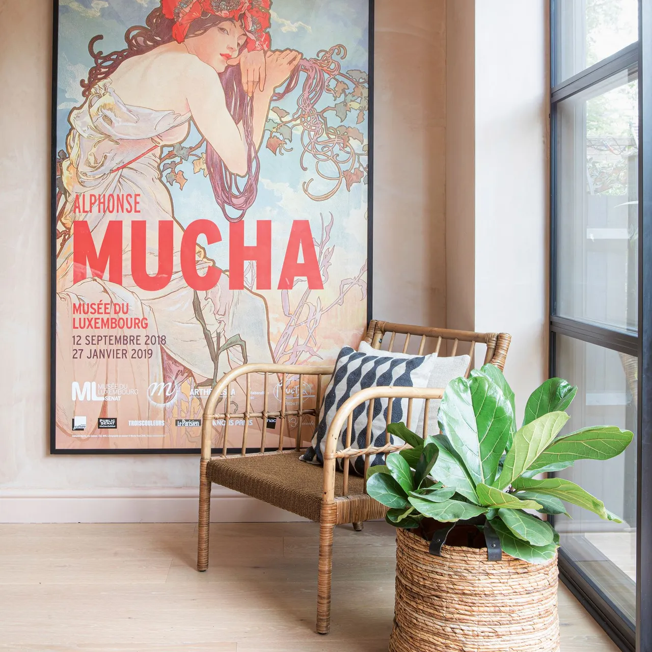 A woven armchair sits beside a large window, a leafy plant and graphic artwork.