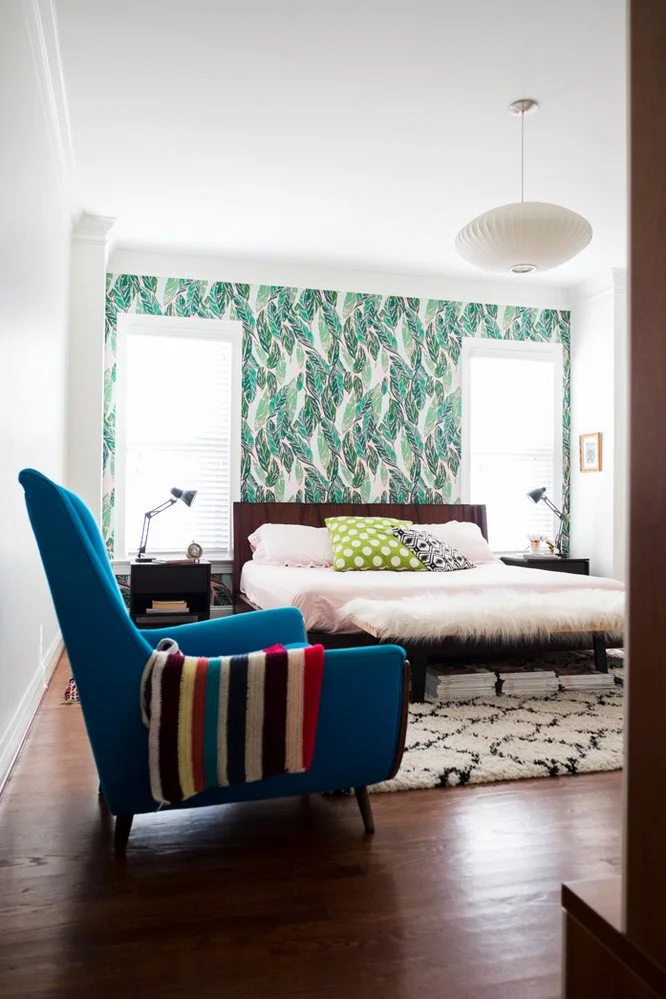 A bedroom with green leafy wallpaper between two tall windows has a dark headboard, pale pink bedding and a blue armchair
