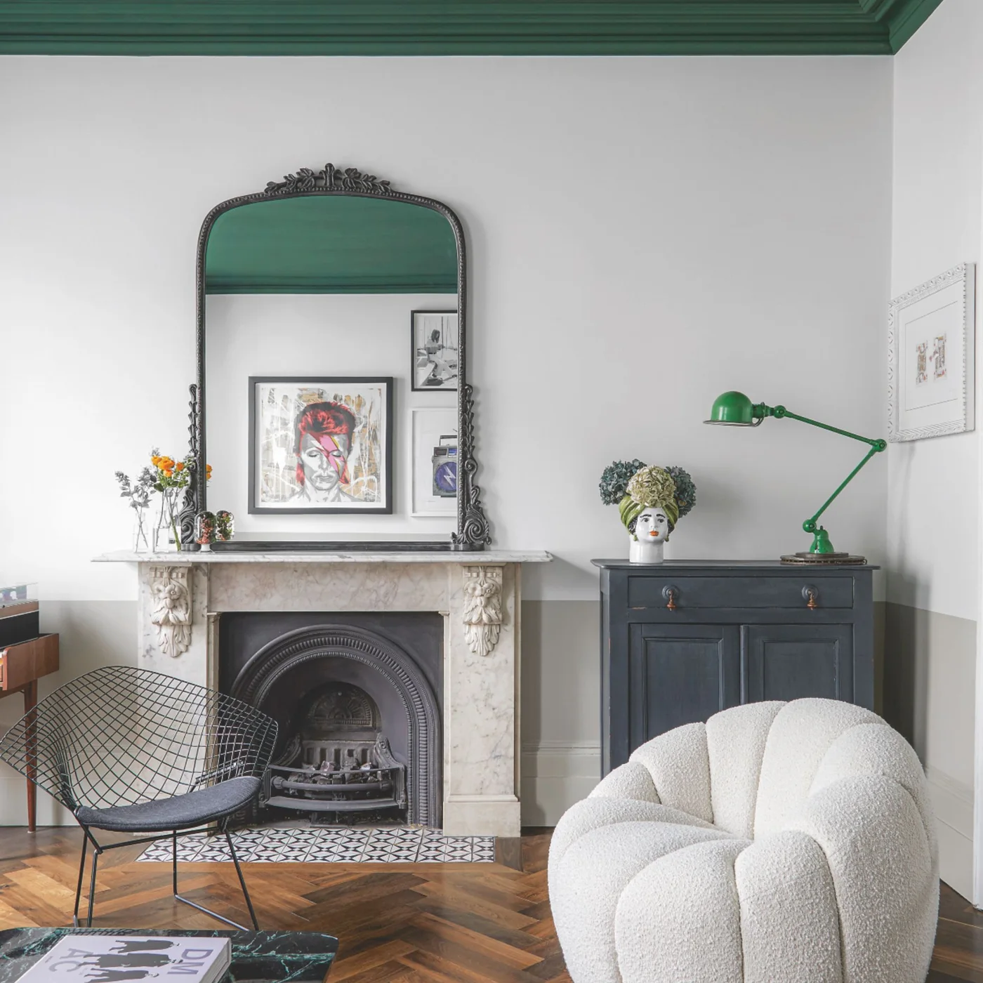 An ornate mirror stands above a pale fireplace beside a wire chair, rounded pale armchair and green lamp