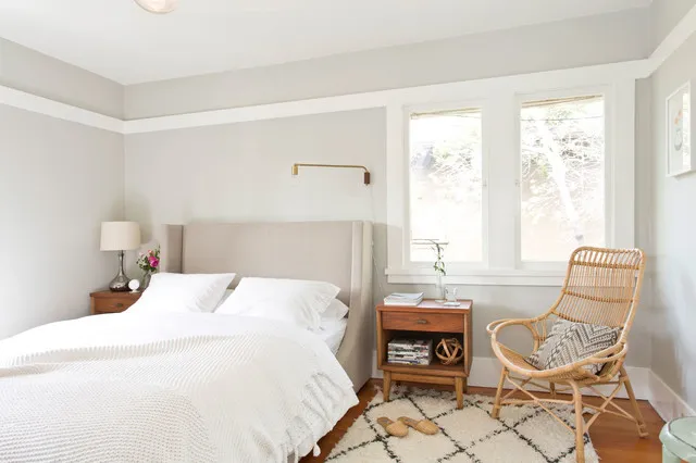 A pale bed sits beside a timber bedside table and open woven chair.