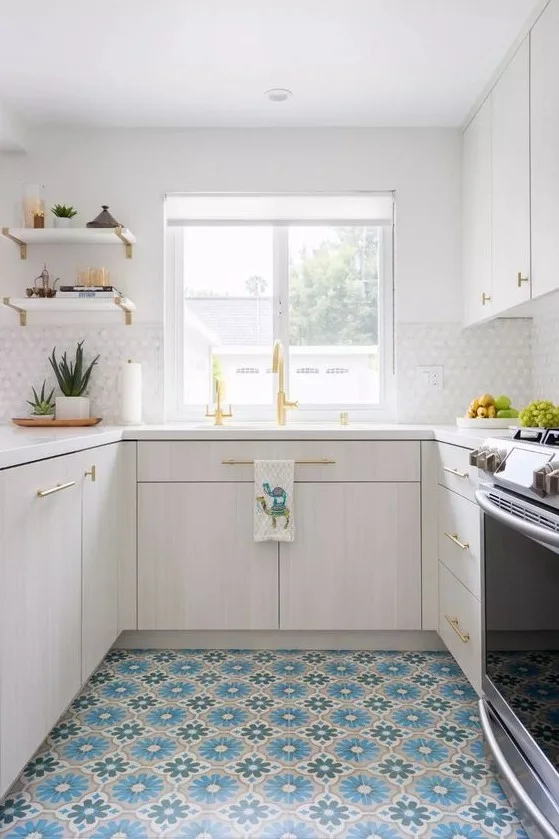 A pale kitchen has white cabinets, light counters and warm-colored sink fittings above a blue, teal and cream floral-patterned floor