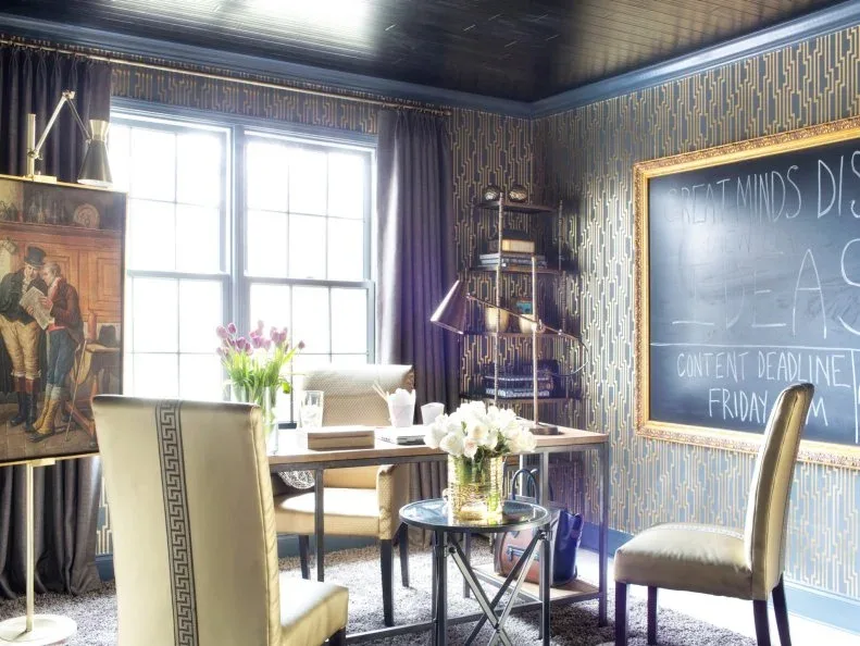 Home office with gold-lined geometric wallpaper, a framed chalkboard, pale chairs and a wooden desk.