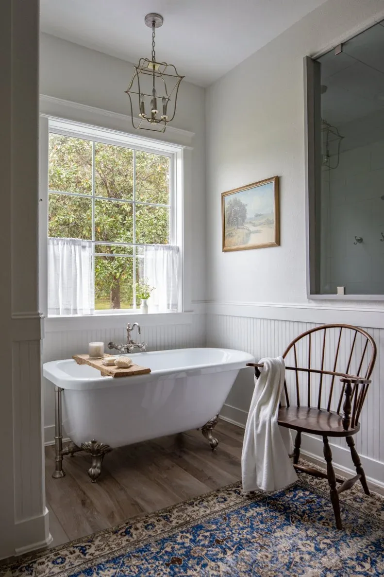 Pale paneled bathroom with white clawfoot tub, dark spindle chair, blue patterned rug, cafe curtain and hanging lantern
