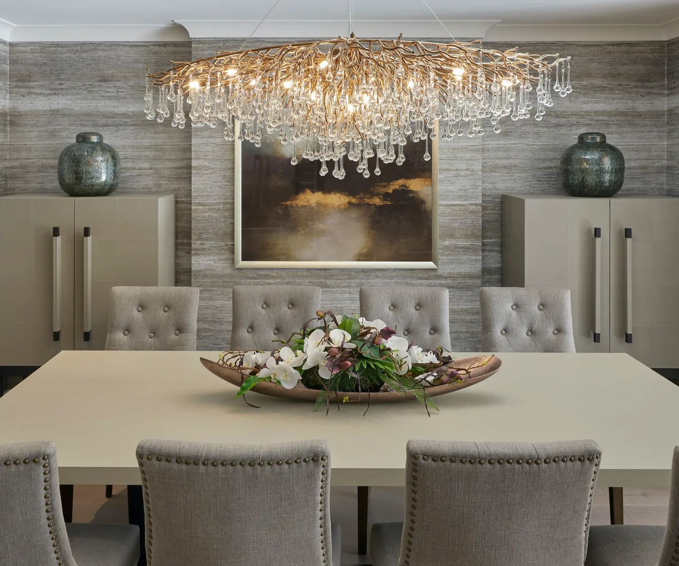 Tufted gray chairs around a pale rectangular dining table beneath a gold-toned branching chandelier with clear drops.