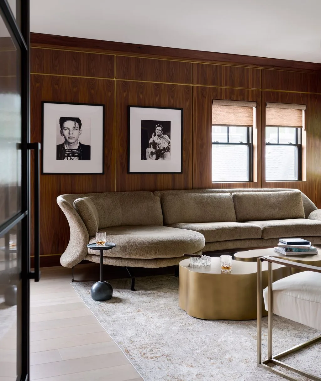 Wood-paneled living room with a curved taupe sofa, two framed monochrome photographs, a pale rug and small gold-colored tables.