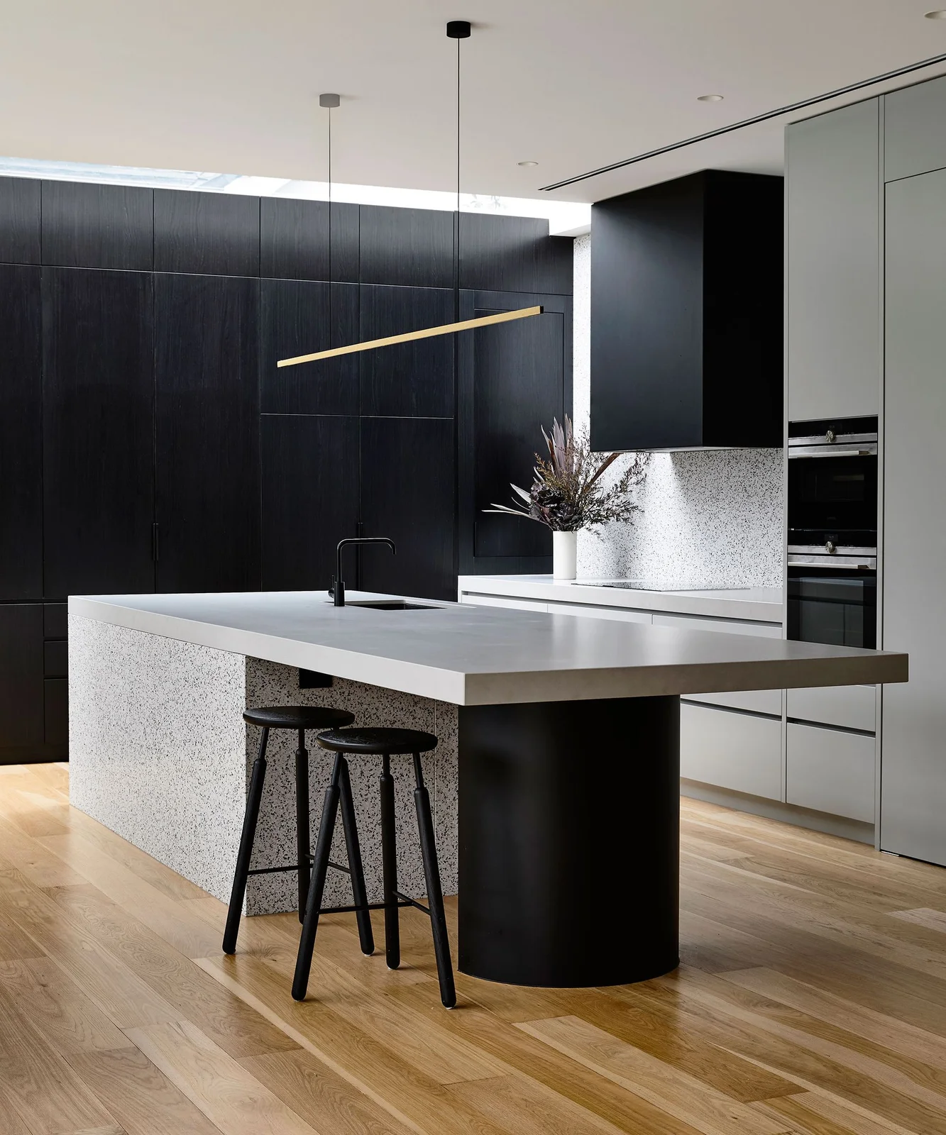 A pale kitchen island top rests above a dark rounded support beside dark stools, tall black cabinets and a slim warm-colored hanging light