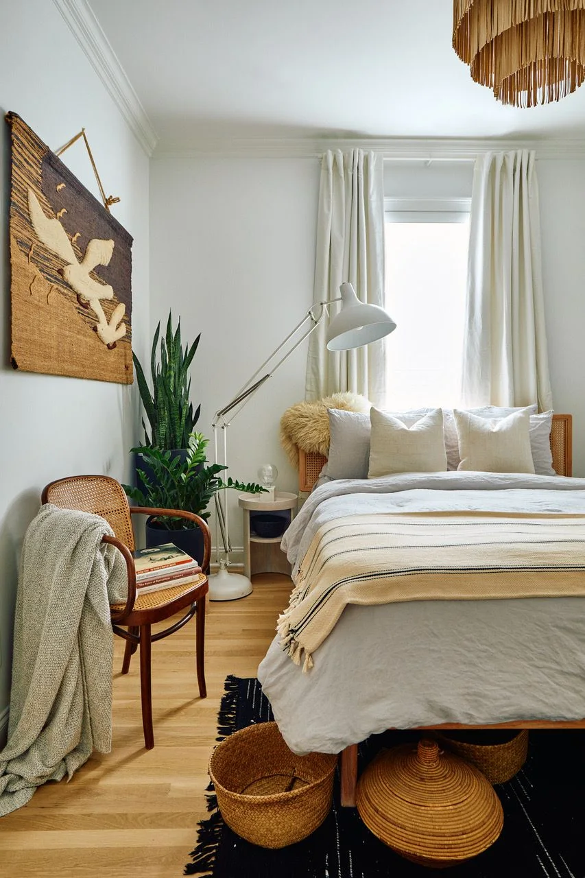 A pale bed with striped layered bedding stands beside a wood-framed woven chair, tall plants and baskets under a fringed pendant near light curtains