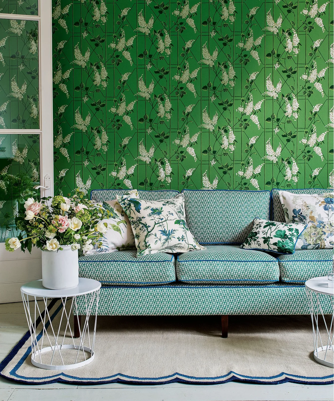 A blue-green patterned sofa with floral cushions sits below bright green floral wallpaper beside a pale side table