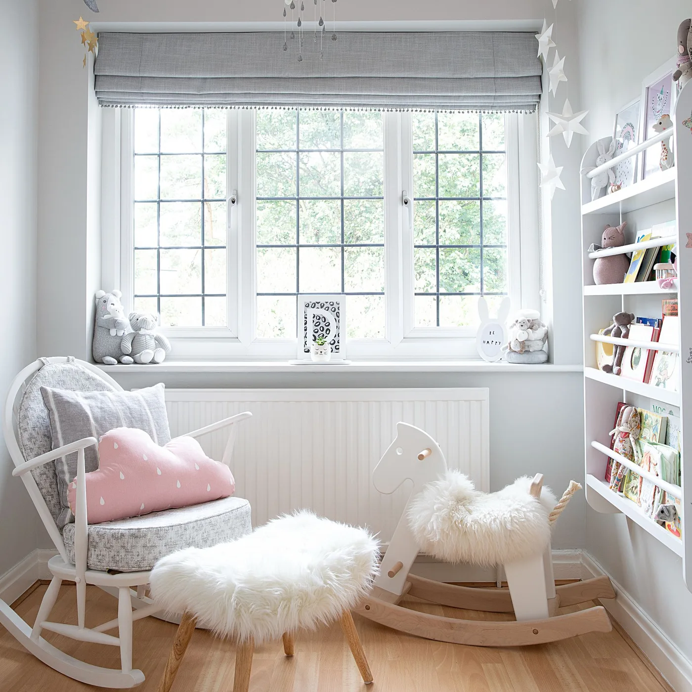 A white and wooden rocking chair with gray upholstery sits beside a window, a book display and a small pale stool