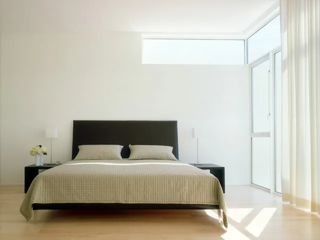 A simple dark headboard stands against pale walls beside high windows.