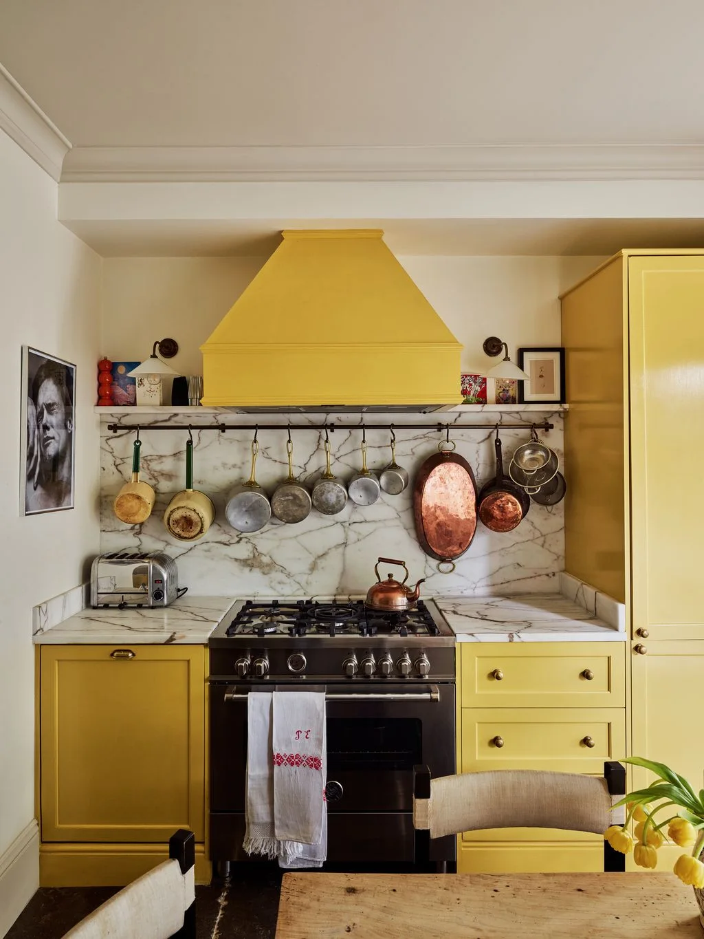 Yellow kitchen cabinets and hood stand above a veined backsplash with assorted hanging pans, framed art and small pieces on a shelf
