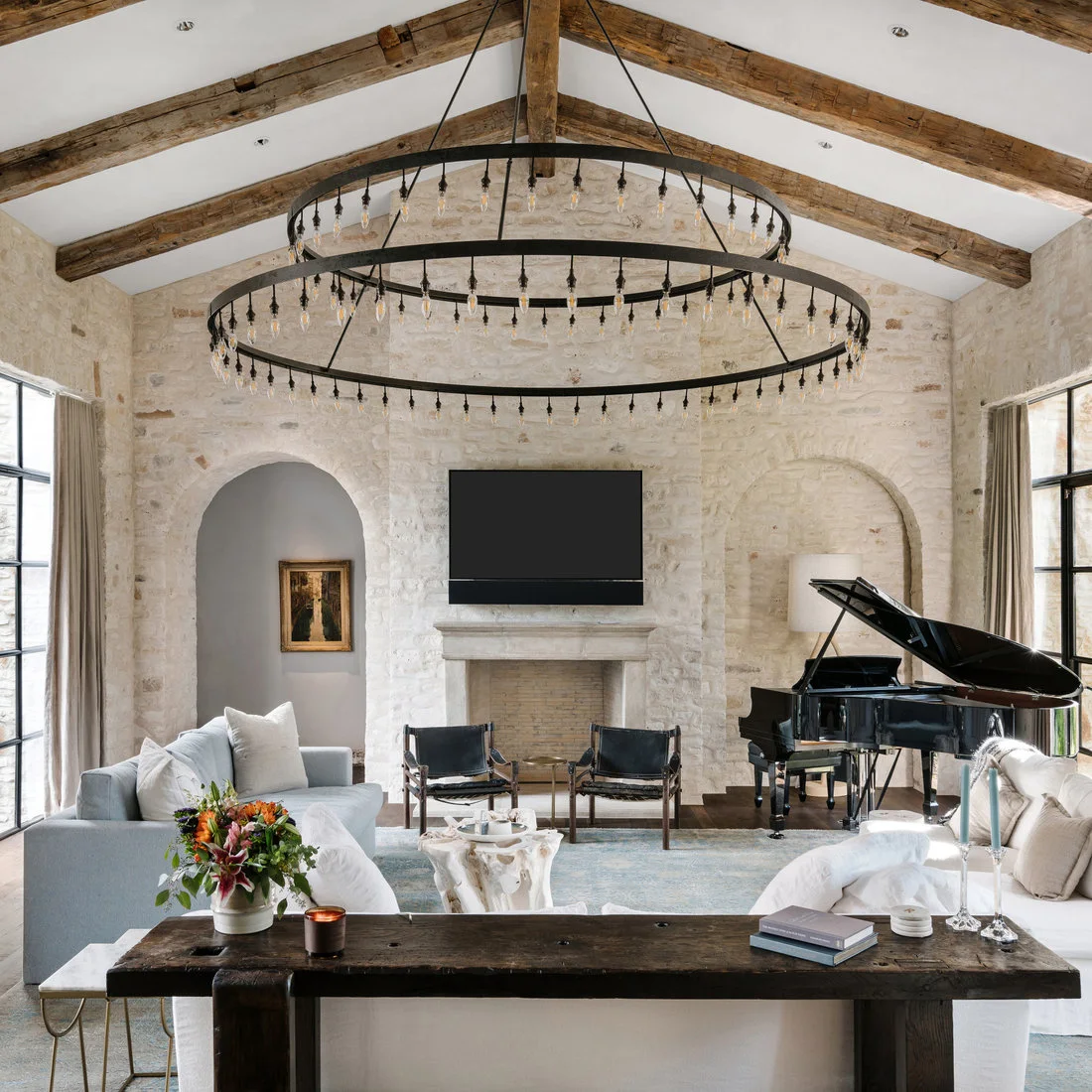 Pale living-room seating surrounds a low table beneath wood ceiling beams and two dark ring lights beside arched wall niches, a fireplace and black piano