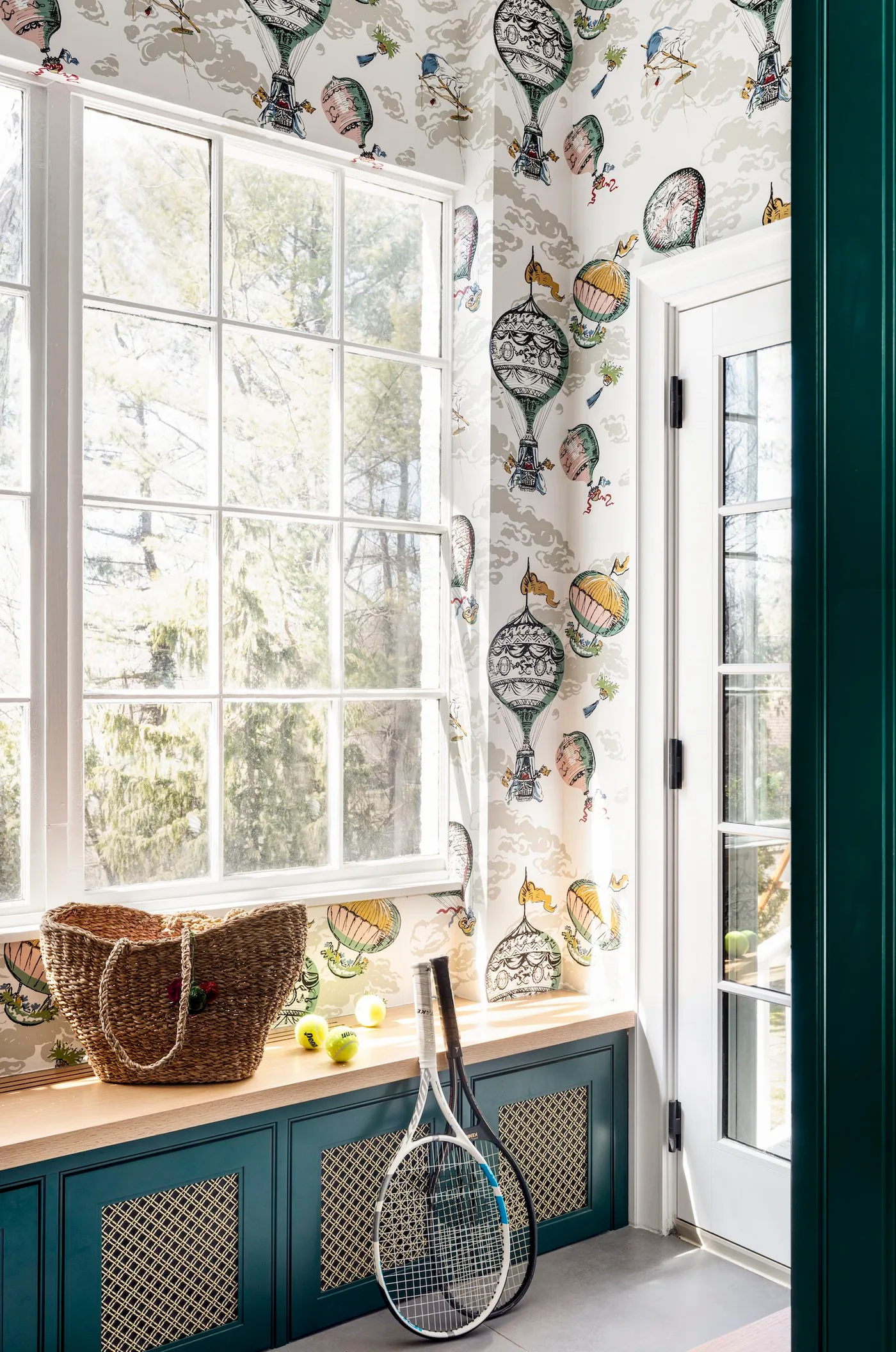 Deep teal lower mudroom cabinets with mesh inserts, wood bench top, a large window, hot-air-balloon wallpaper, woven basket, and tennis rackets.