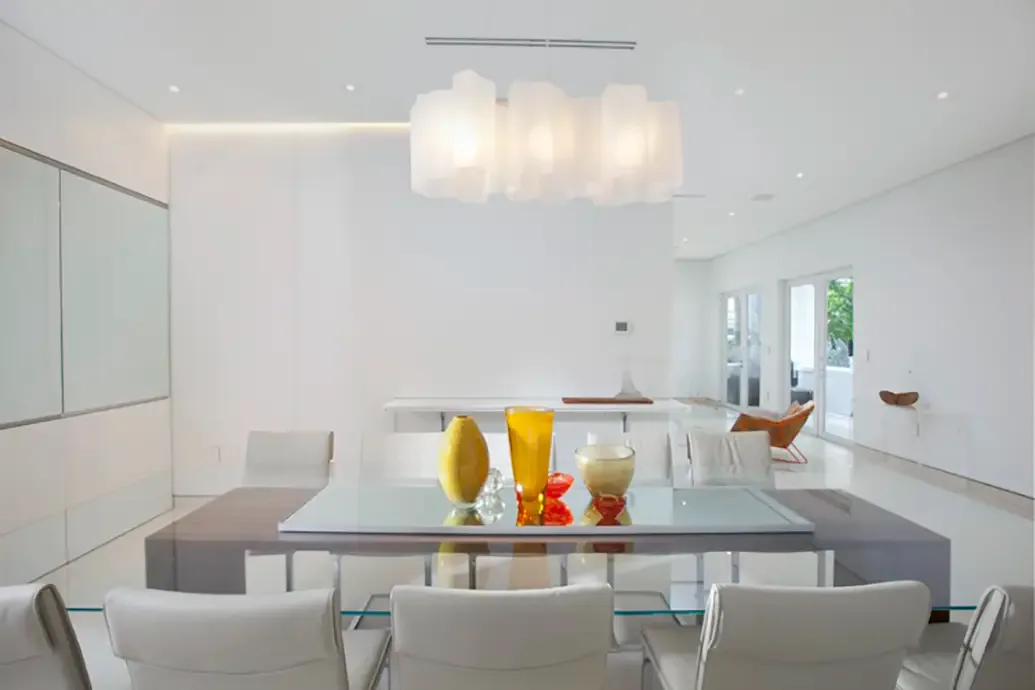 A clear glass dining table with white chairs and gray benches stands in a pale room below white hanging lights with small yellow and orange vases