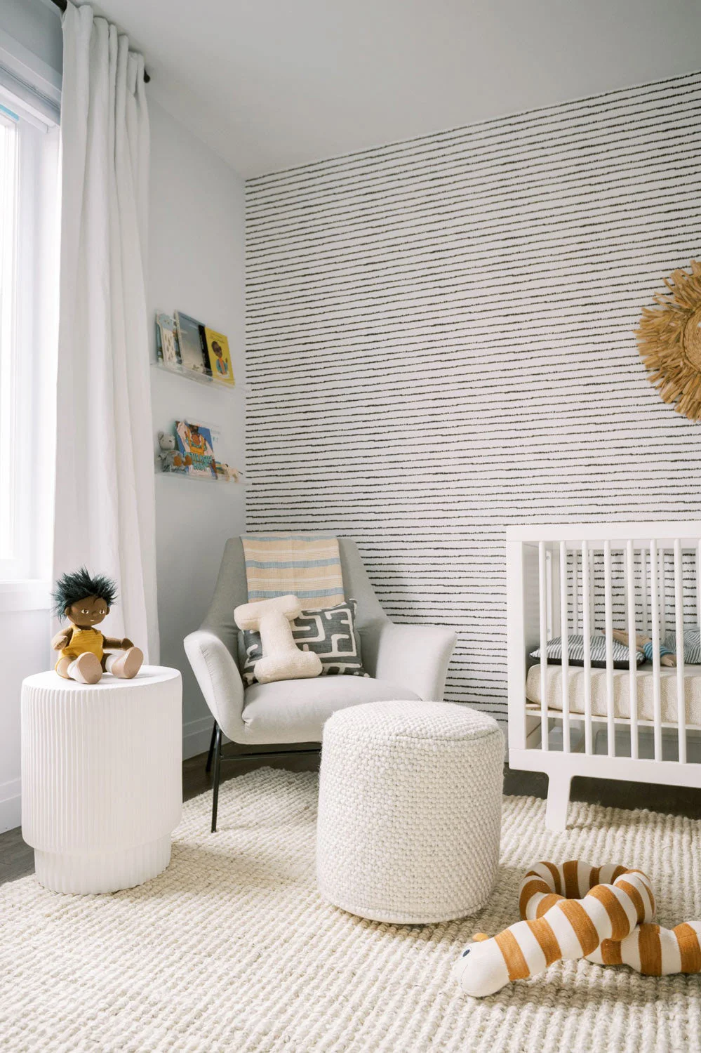A pale nursery adult chair with gray patterned cushion sits beside a white cylindrical table and textured footrest below fine dark wall lines