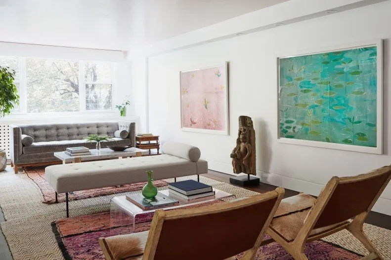 Living room with narrow pale upholstered bench, low clear table holding books, wooden chairs, gray tufted sofa, pink and green artwork and standing wooden sculpture.