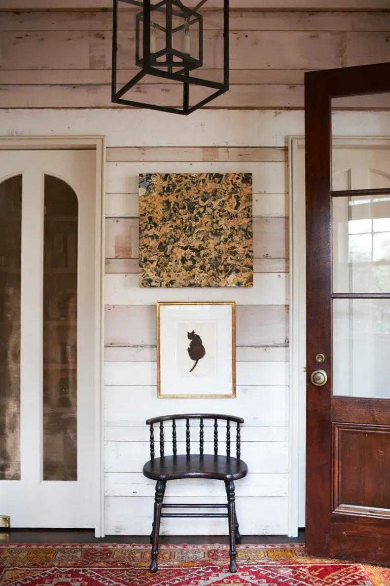 White horizontal-board entry with dark wood door, black spindle chair, two framed artworks, red patterned rug and dark cage lantern