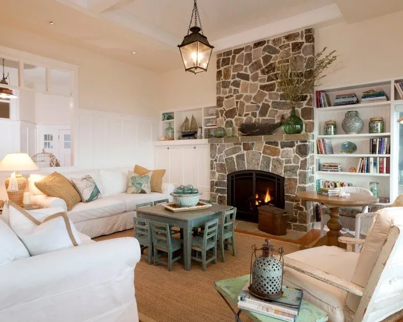 Pale country living room with stone fireplace, white shelving, light sofas, chairs and a small green game table at the center