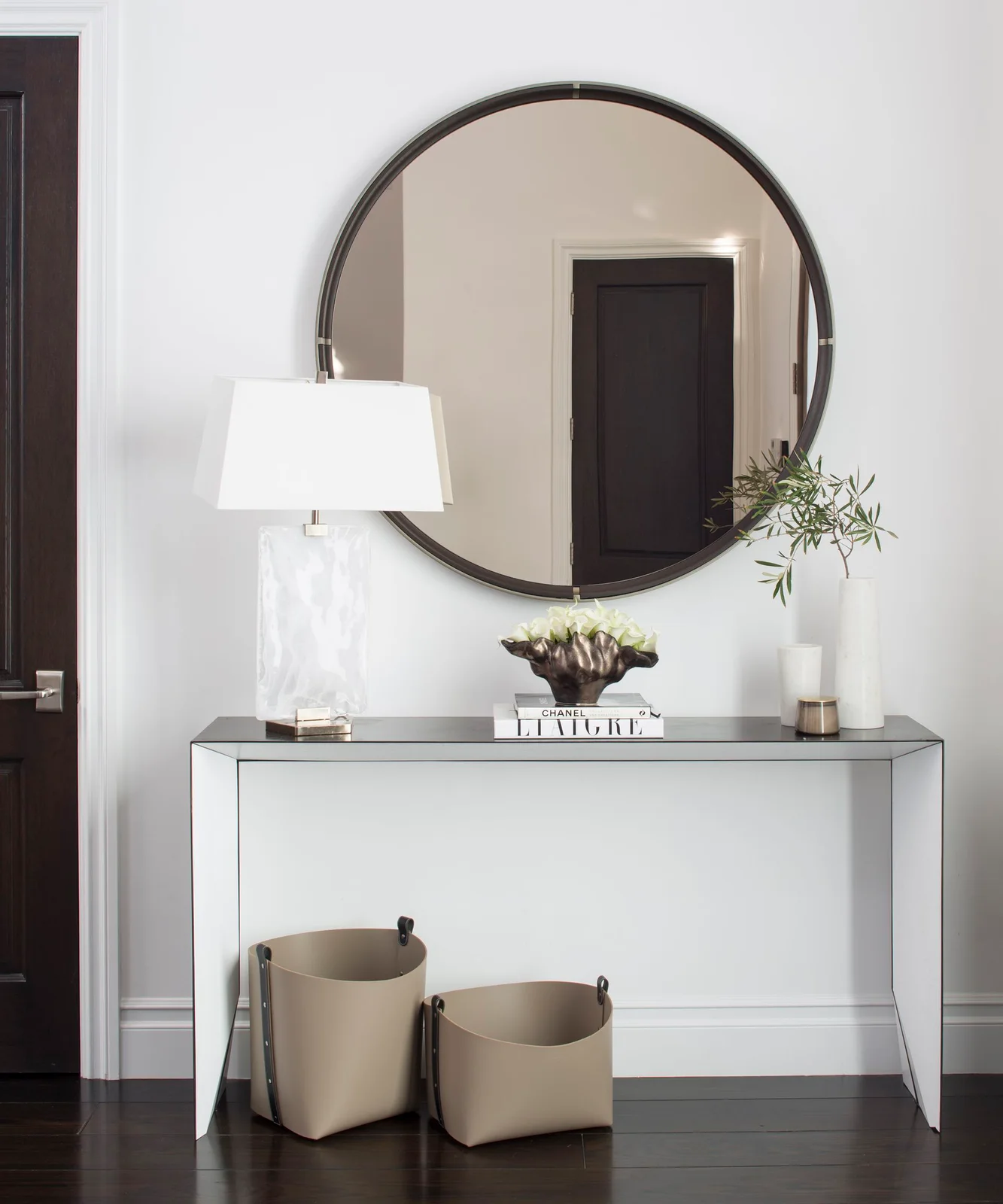 A pale console with darker top and thin edges stands below a large dark round mirror beside a white lamp, pale walls and dark flooring