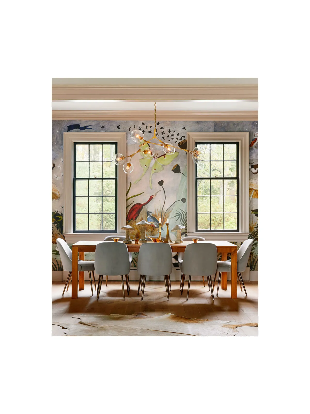 A colorful scenic wall frames a timber table between tall windows.