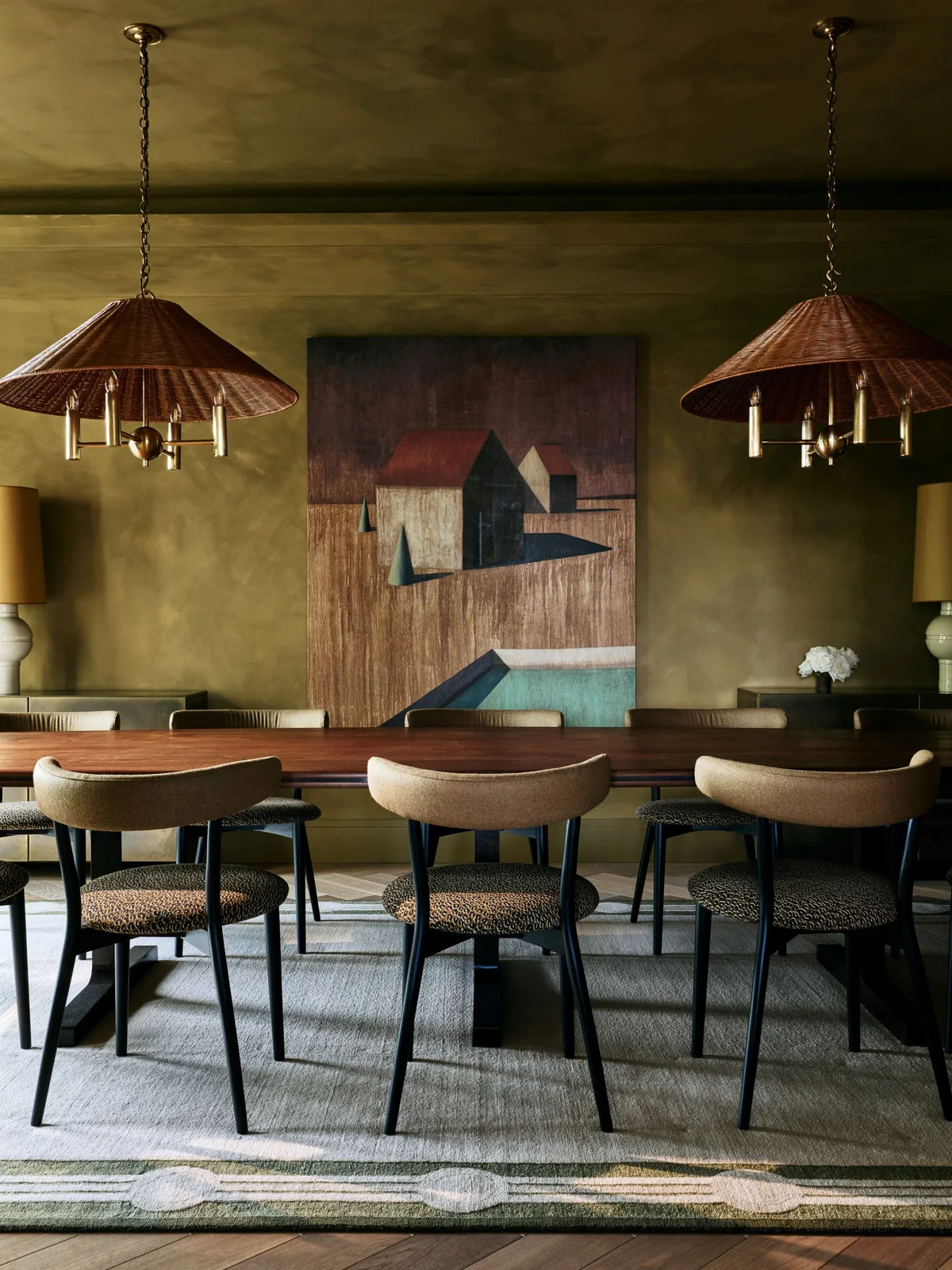 Olive-toned walls surround a long wooden dining table, curved upholstered chairs, two wide woven-looking pendants and large geometric artwork