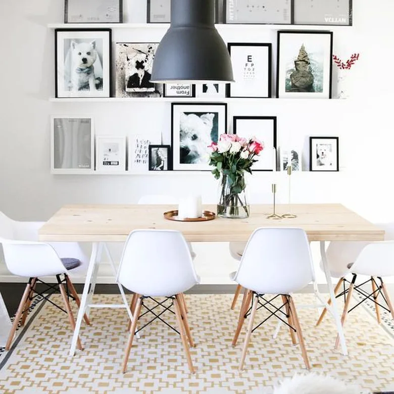 White picture ledges filled with framed prints behind a pale dining table, white chairs and a black pendant